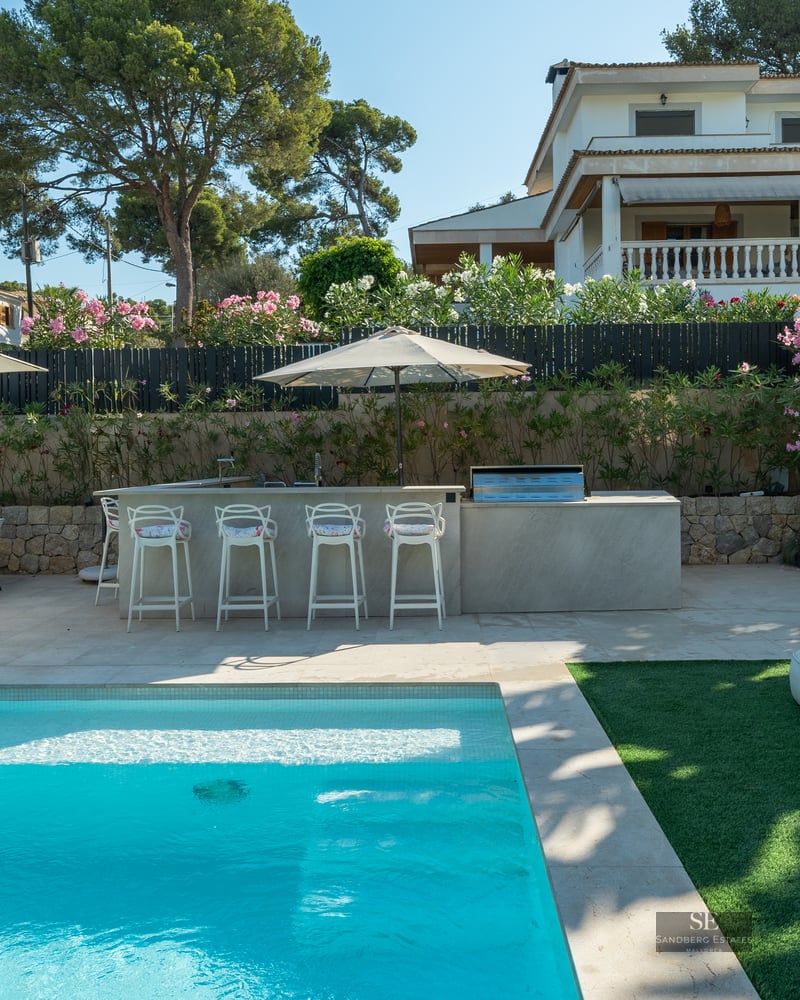 Bright swimming pool area with modern outdoor kitchen, bar stools, and dining set under umbrellas in a sunny garden.