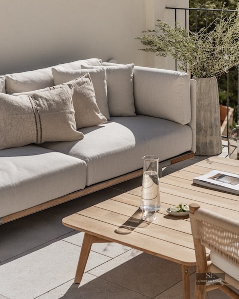 Modern outdoor terrace featuring a plush grey sofa, wooden coffee table, and greenery against a white wall and railing.