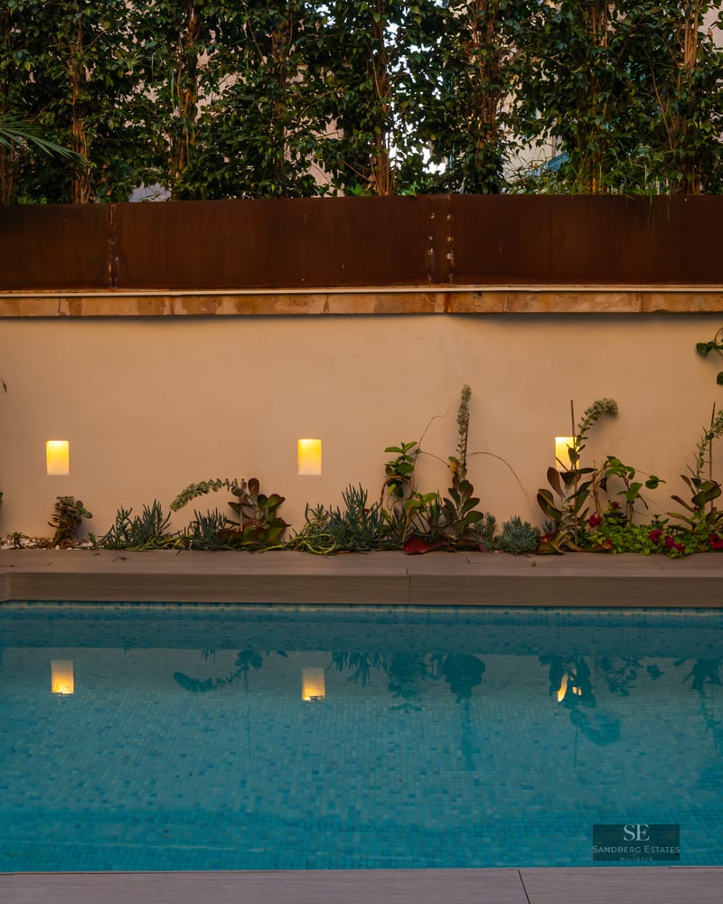 Small rectangular blue-tiled pool at dusk with warm wall lights, wood-effect decking, and lush greenery.