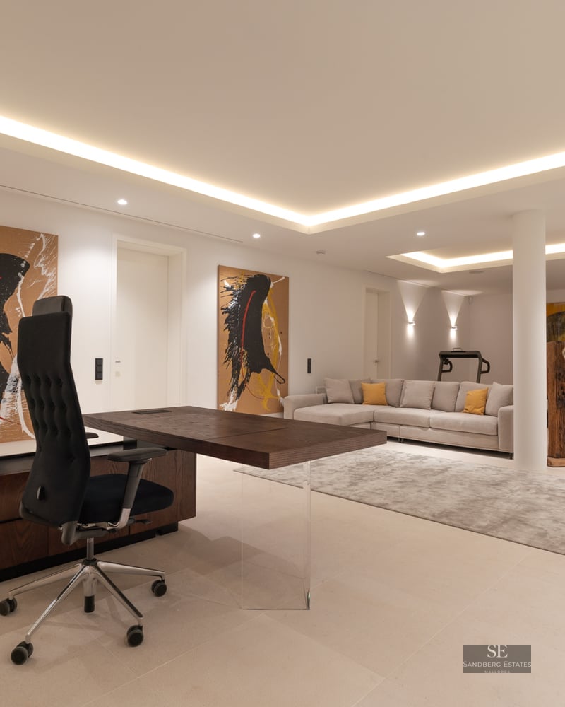 Spacious modern home office with a dark wood desk, grey sofa, abstract art, and gym equipment in the background.