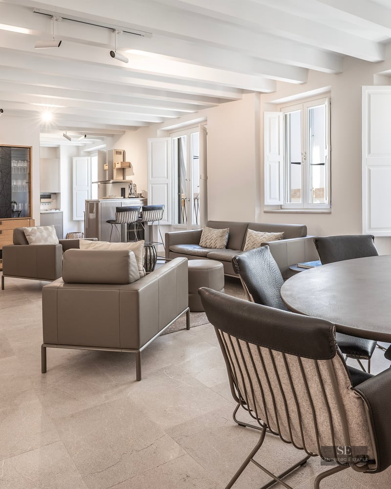 Salon spacieux avec plafonds à poutres blanches, meubles en cuir gris et table à manger ronde.