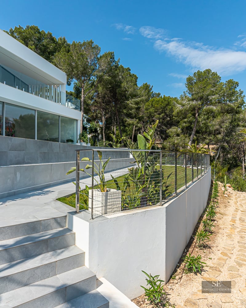 Modern white villa with a tiered garden featuring stone stairs, a lawn, and palm trees under a clear blue sky.