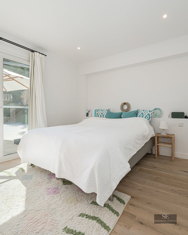 Chambre lumineuse avec un grand lit, parquet et portes vitrées s'ouvrant sur une piscine turquoise et un parasol.