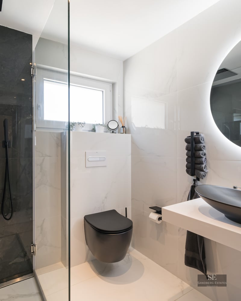Salle de bain de luxe avec douche en marbre noir, murs en marbre blanc, vasque noir mat et miroir circulaire rétroéclairé.