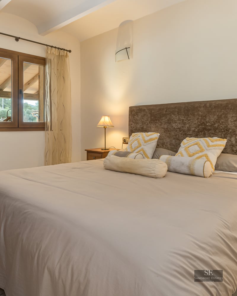 A cozy bedroom featuring a large bed with a brown velvet headboard, wooden nightstands, and warm ambient lamp lighting.