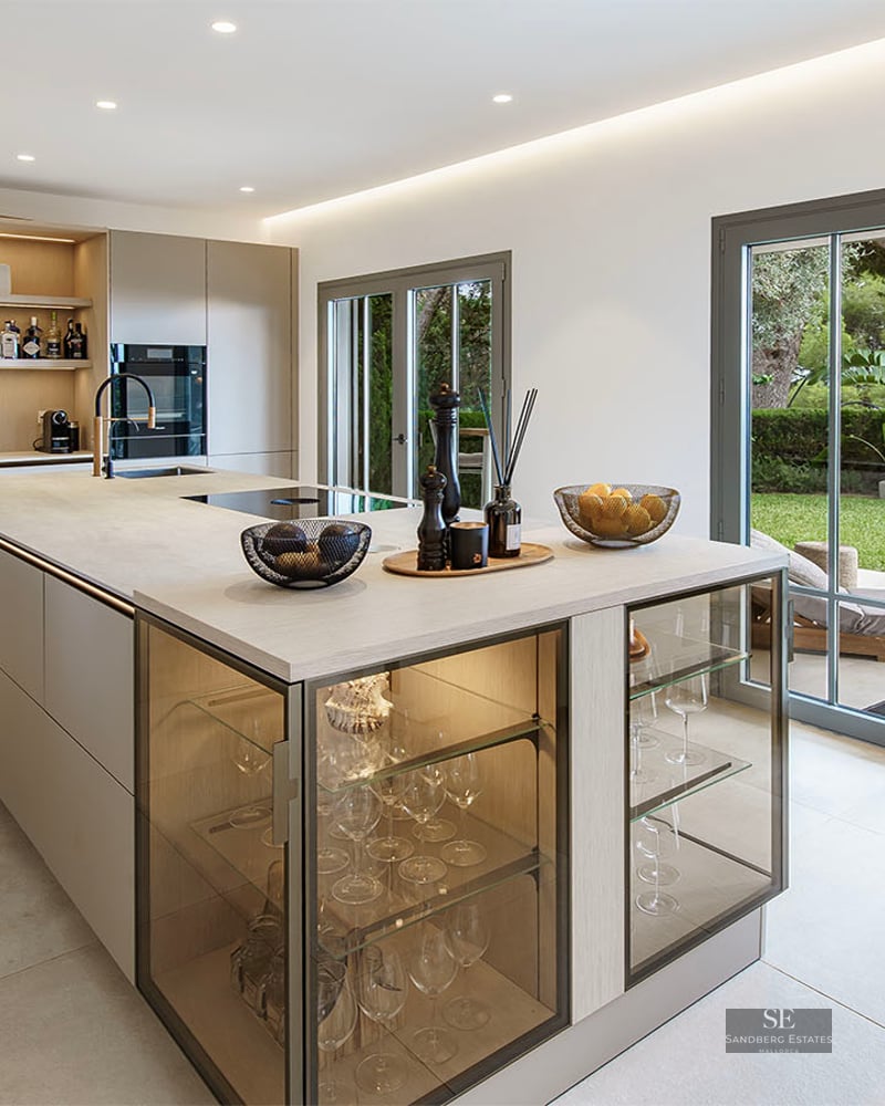Modern kitchen featuring a large island with integrated glass displays and glass doors opening to a green garden.