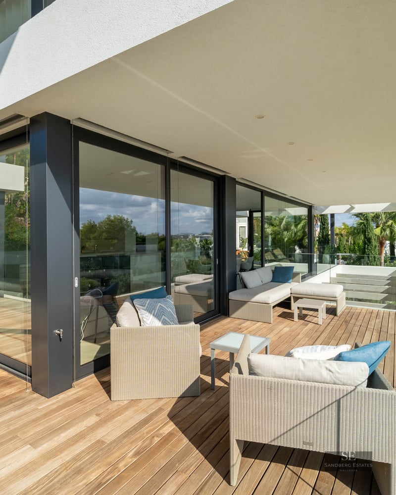 Spacious wooden terrace with designer lounge furniture and glass railings overlooking a swimming pool and lush garden.