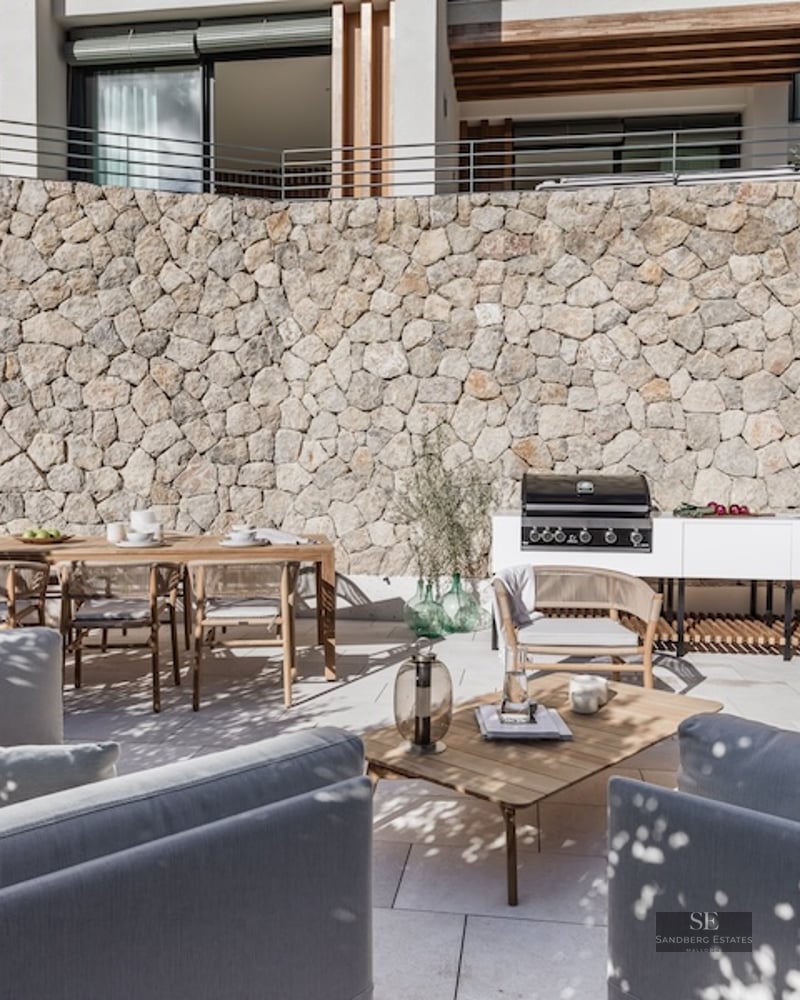 Modern terrace with a stone wall, outdoor kitchen, wooden dining table, and grey sofas under dappled natural light.