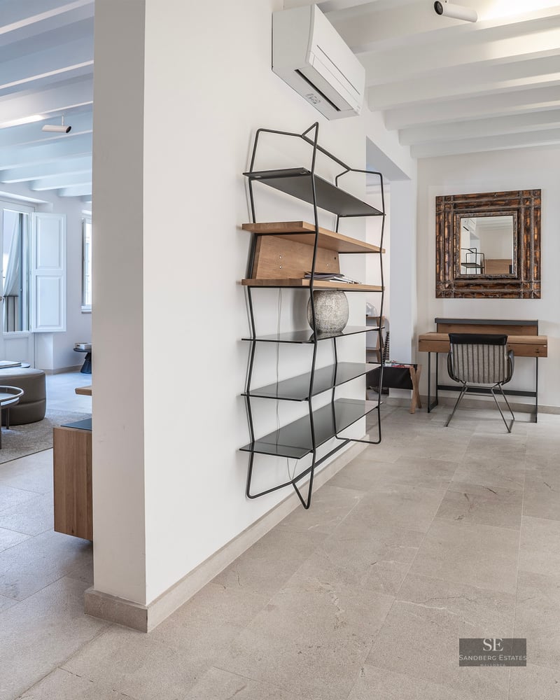 Grand salon moderne avec canapé en cuir gris, sols en pierre, poutres apparentes et étagère minimaliste.