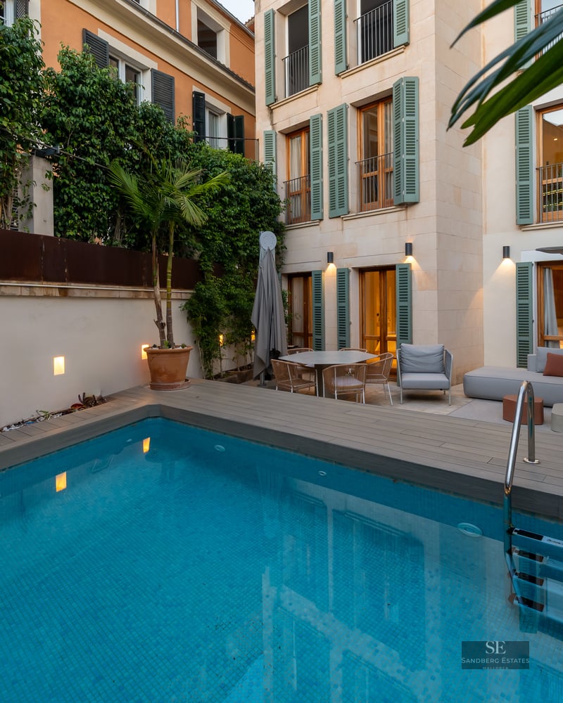 Modern private pool area with wooden decking, grey outdoor sofas, and a Mediterranean-style building with green shutters.