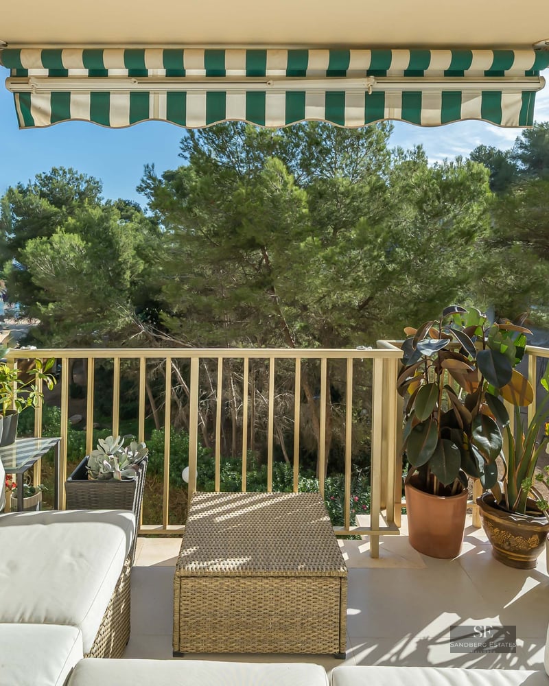 Balcon lumineux avec canapés blancs, table en osier, store rayé et plantes donnant sur les arbres et les bâtiments au loin.