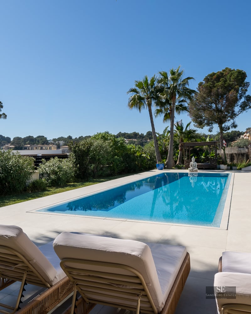 Turquoise swimming pool surrounded by white stone tiles, sun loungers, palm trees, and a Mediterranean villa under a blue sky.
