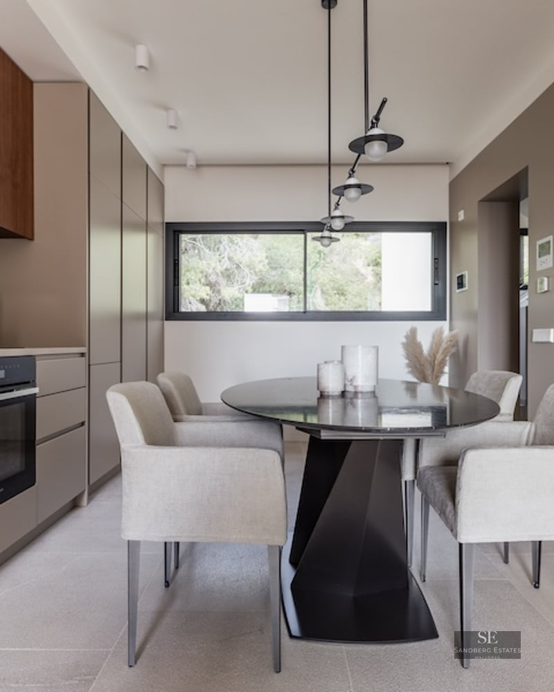 Modern dining room with oval black table, grey armchairs, and taupe cabinetry under designer pendant lights.