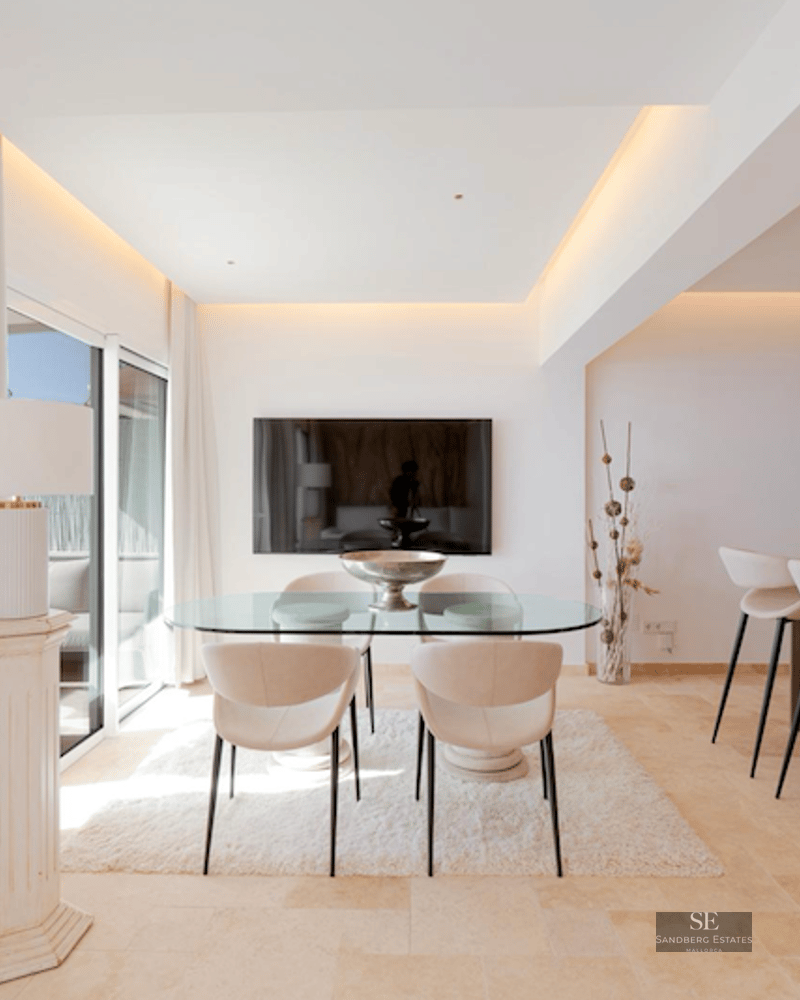 Minimalist dining room featuring a glass table, cream chairs, white rug, and a breakfast bar in the background.