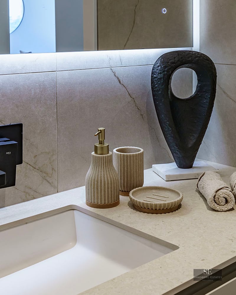 Close-up of a white sink with a matte black wall-mounted faucet, beige marble-effect tiling, and designer accessories.