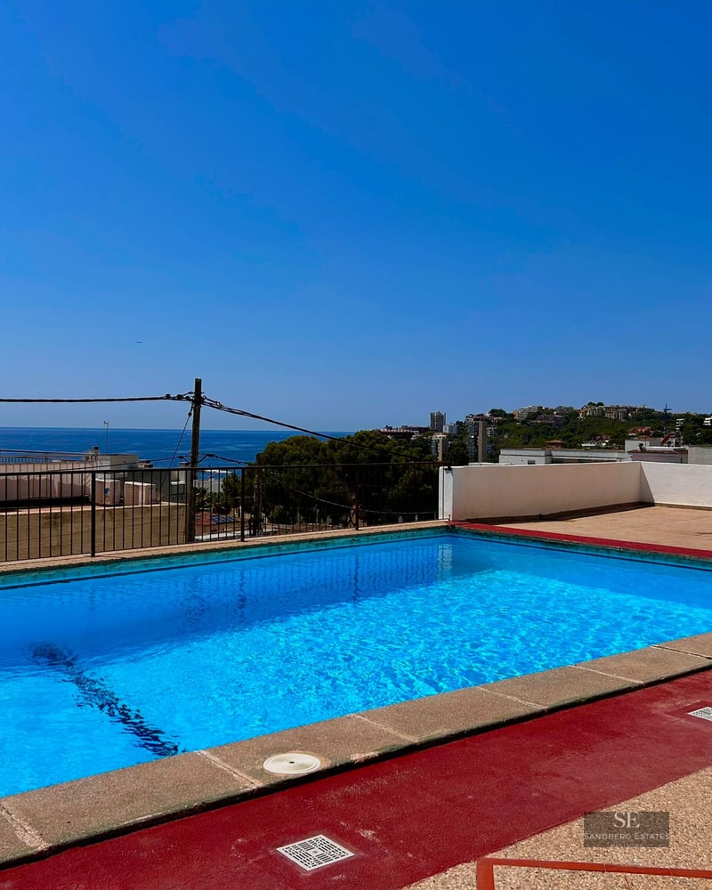 Blue rooftop pool with red deck overlooking the Mediterranean sea and coastline under a clear blue sky.