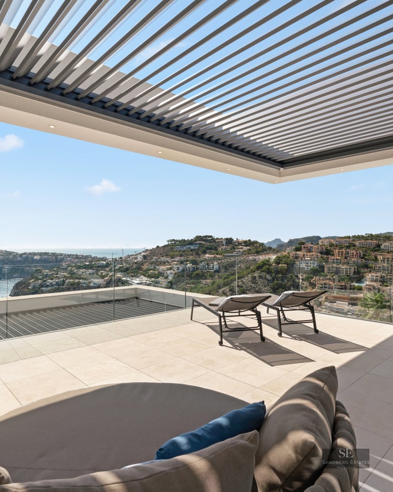 Modern terrace with stone tiling, sun loungers, a louvered pergola, and stunning views of the sea and coastal hills.