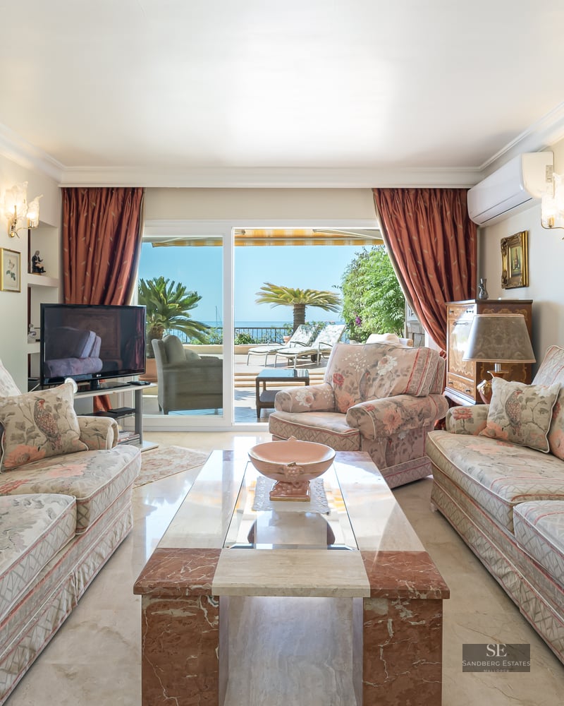 Spacious living room with floral sofas, marble table, and large glass doors opening to a terrace overlooking the sea.