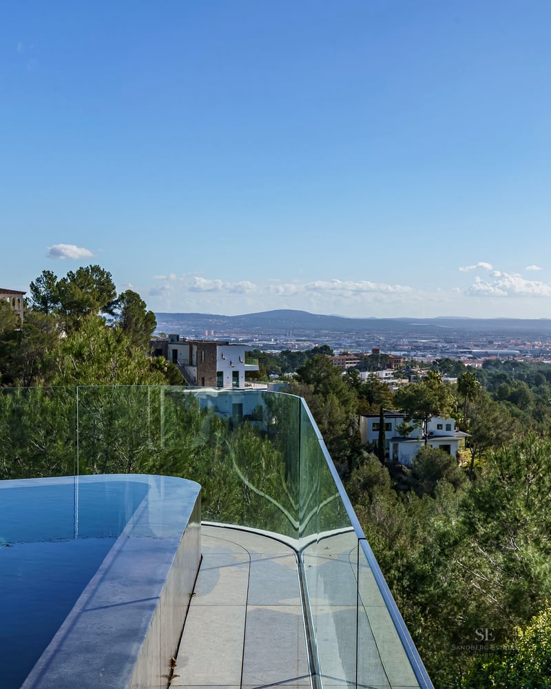 An infinity-edge pool with glass railing overlooking a lush forested valley and distant city.