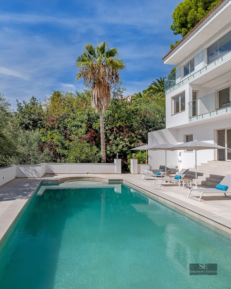 A large turquoise swimming pool next to a modern white villa with lounge chairs, umbrellas, and a Buddha statue.