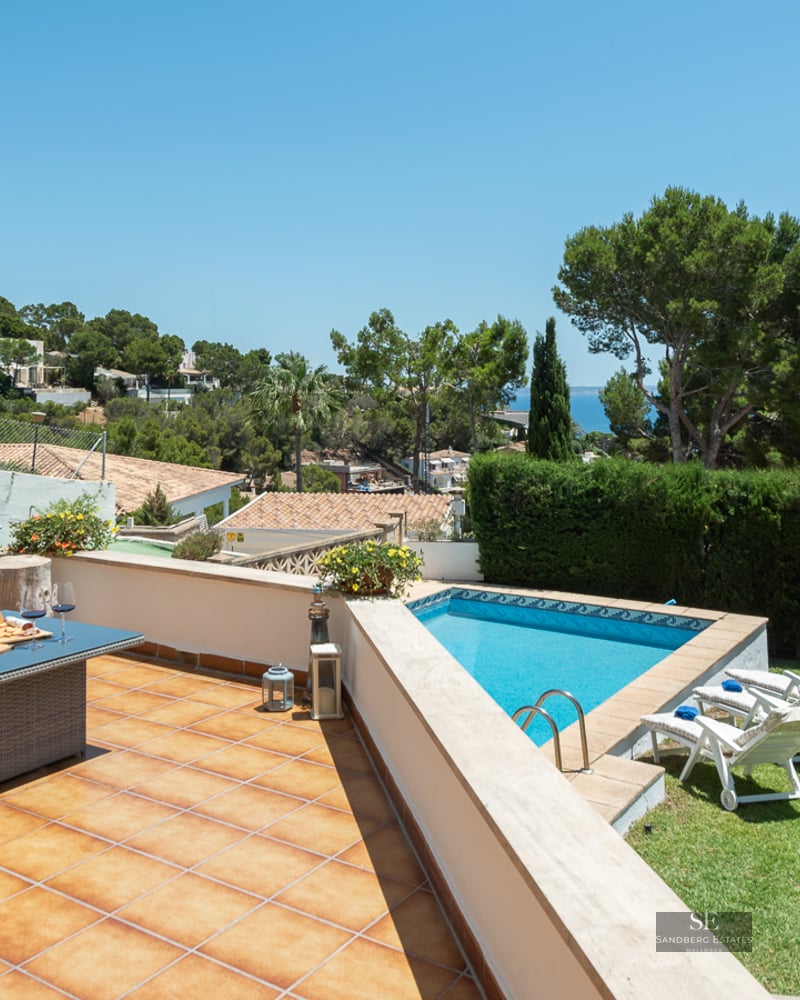 Sun-drenched terrace with grey wicker sofas, a blue swimming pool, green lawn, and panoramic ocean views.