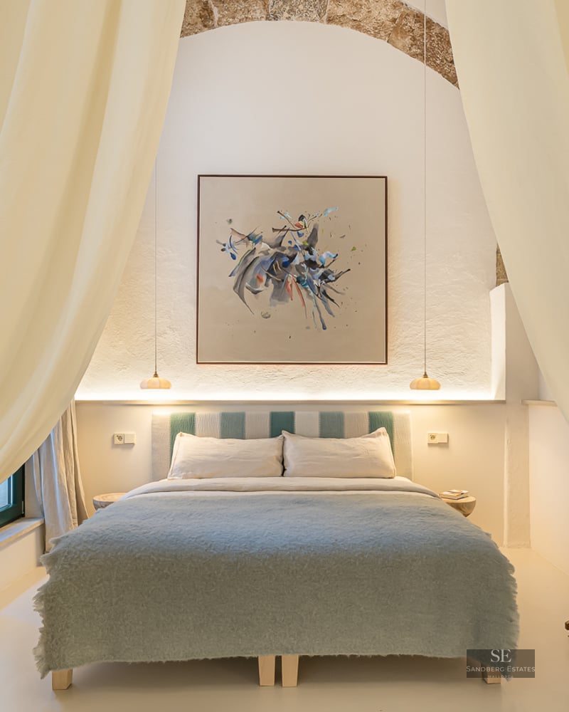 Chambre élégante avec grand lit, rideaux crème, arche en pierre apparente et tableau abstrait moderne.