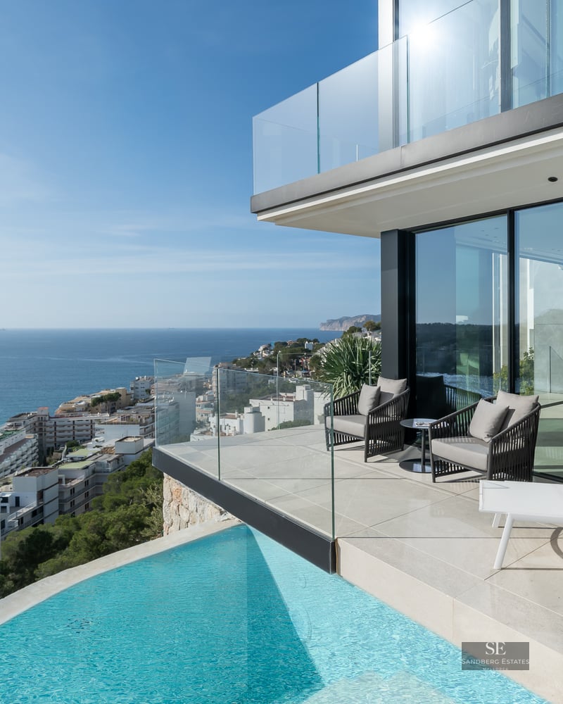 Modern infinity pool and terrace with grey armchairs overlooking the Mediterranean sea and a coastal town.