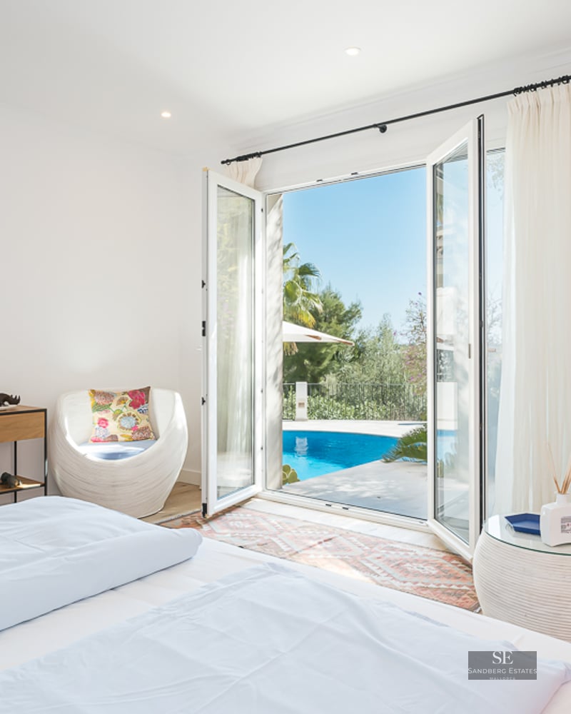 Chambre lumineuse avec lit blanc et chaises design, s'ouvrant sur une piscine turquoise et un jardin.