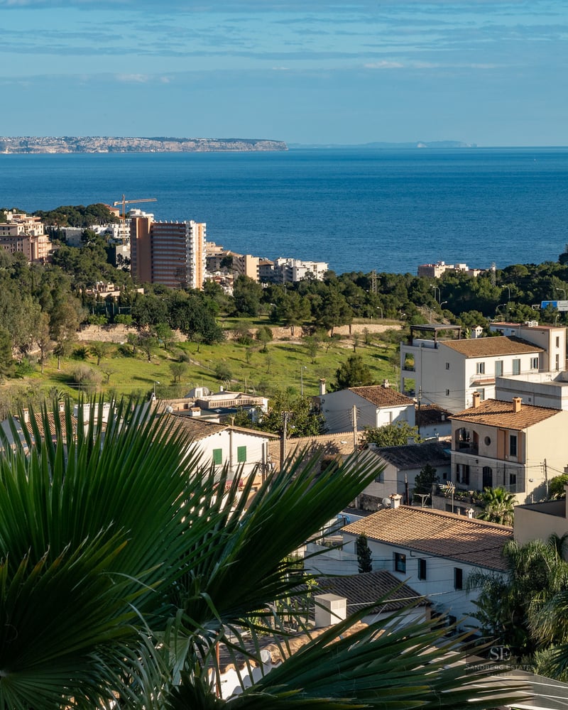 Elevated view of a coastal town with Mediterranean houses, lush greenery, and the blue sea in the distance.