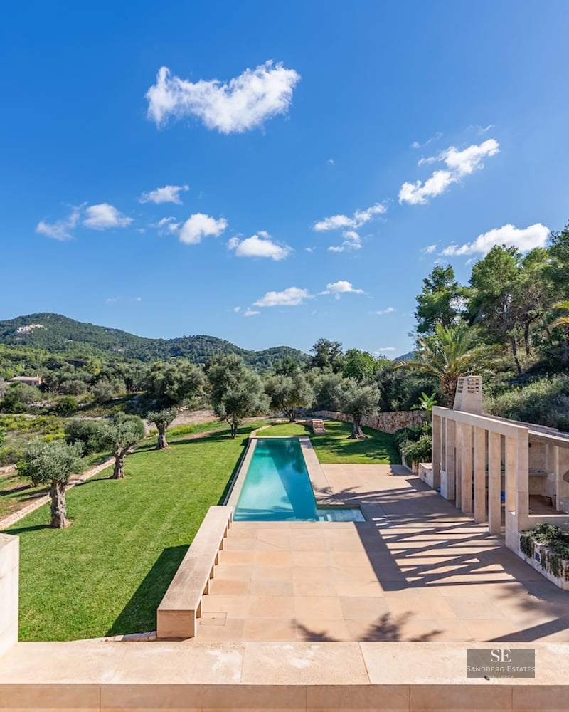 High-angle view of a long turquoise lap pool, stone terrace, and olive trees set against lush green hills and a blue sky.