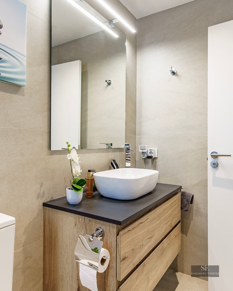 Modern bathroom featuring a white vessel sink on a wooden vanity, beige tiled walls, and a large mirror with LED light.