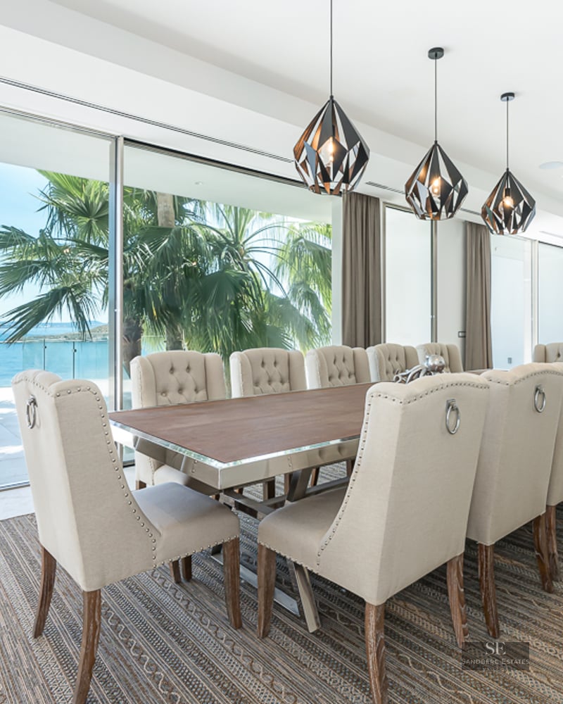 Elegant dining room featuring a long table, beige tufted chairs, and large glass doors opening to a sea view with palm trees.