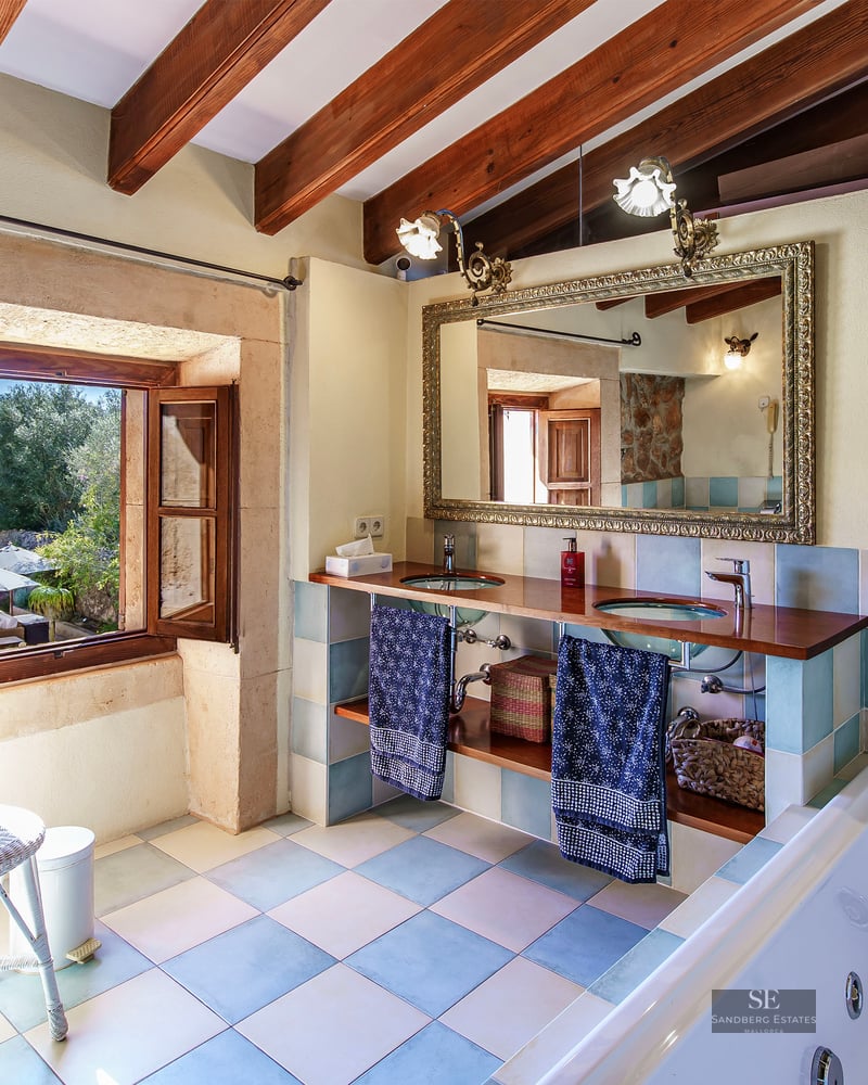 Master bathroom with exposed wood beams, double glass sinks, ornate mirror, and whirlpool tub overlooking a garden.
