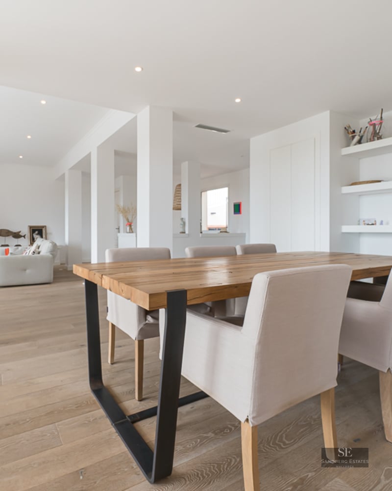 Salle à manger moderne avec grande table en bois et chaises rembourrées, s'ouvrant sur un salon lumineux avec cheminée.