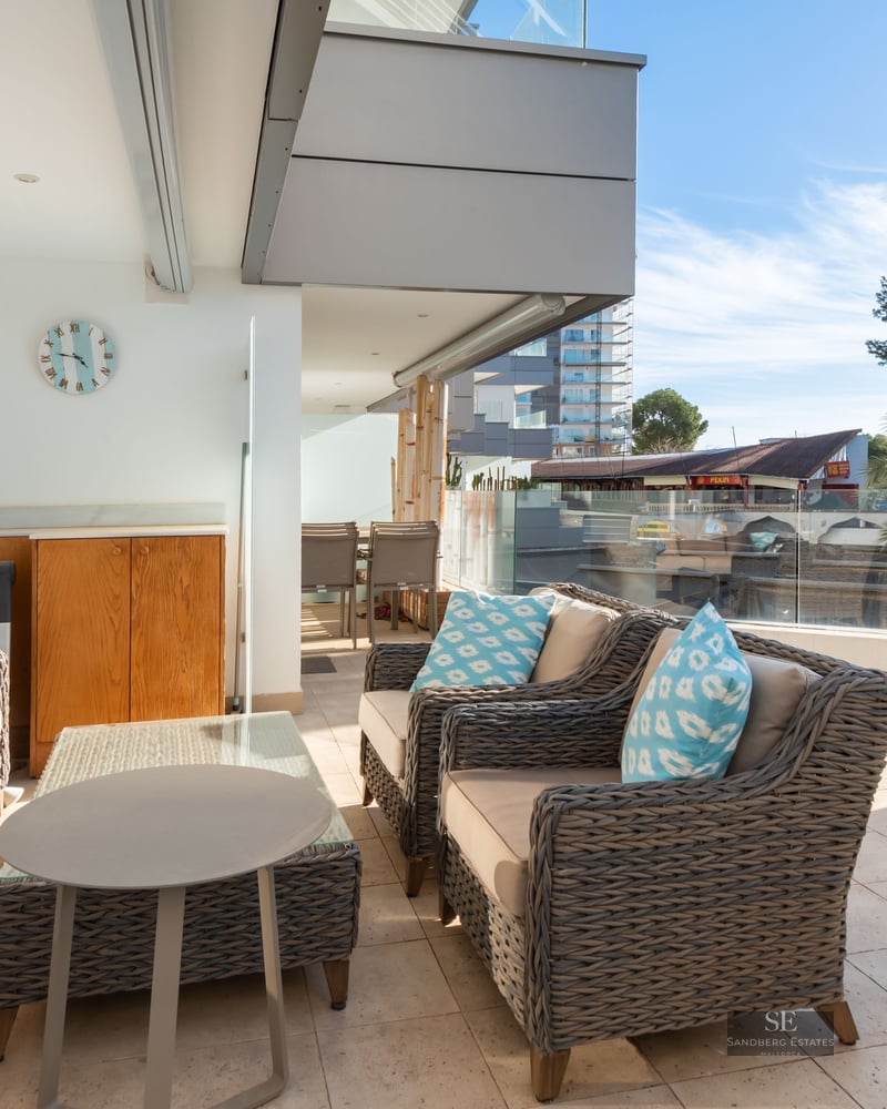 Grande terrasse avec canapés en osier gris, coussins bleus et garde-corps en verre sous un ciel bleu.