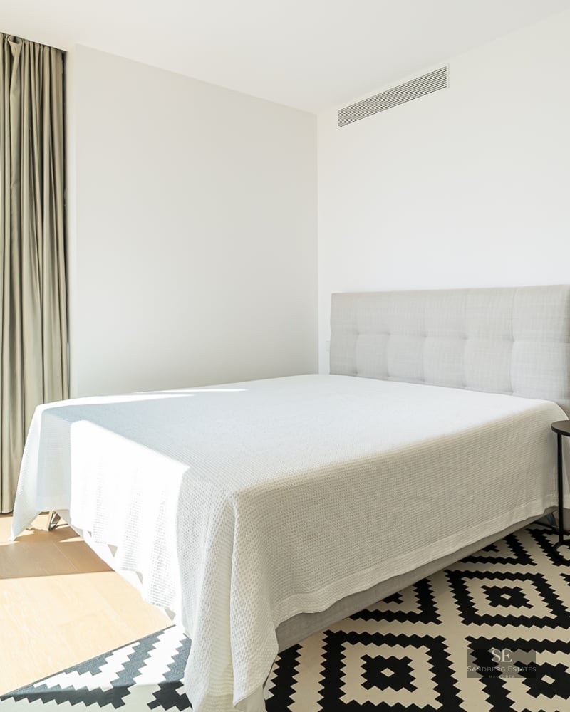 Modern bedroom featuring white bedding, a geometric rug, floor-to-ceiling window, and an ensuite bathroom.
