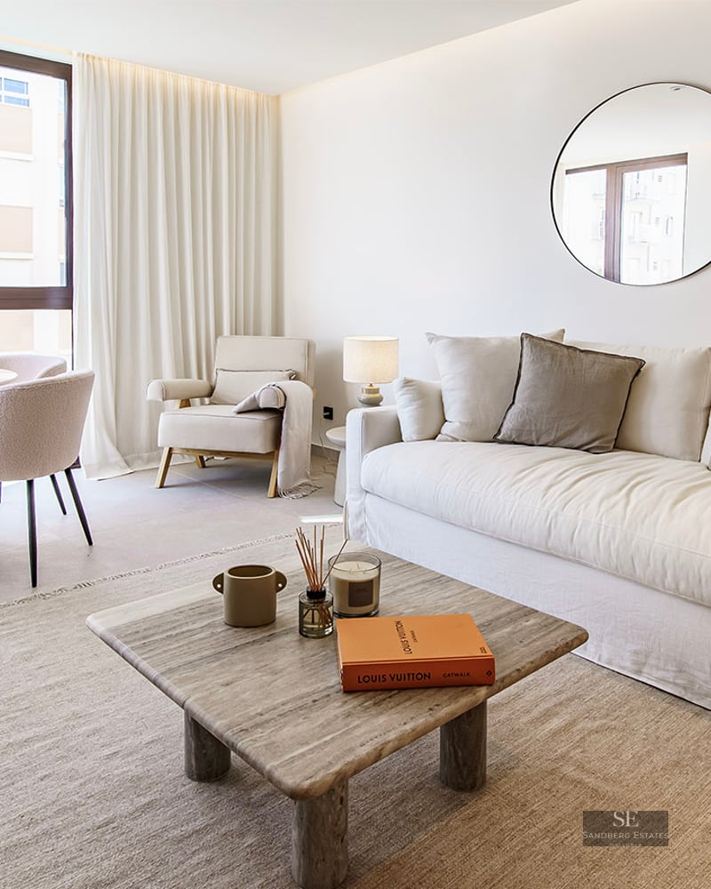 Bright living room featuring a large white sofa, stone coffee table, two round mirrors, and a dining table by a window.