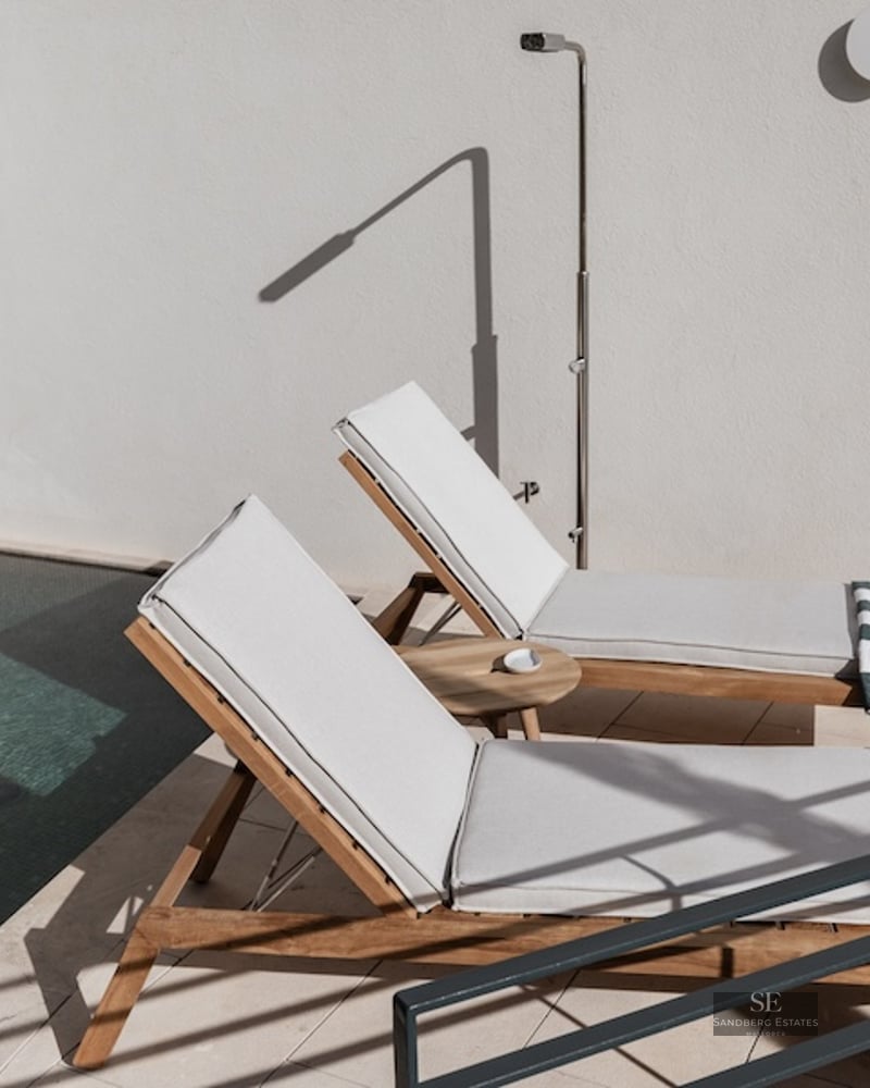 Two wooden sun loungers with white cushions next to a pool and outdoor shower on a sunny terrace.
