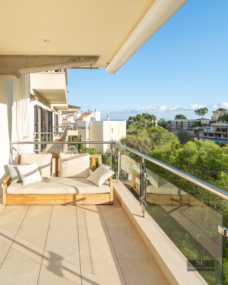 Modern balcony with wooden sofa and glass railing overlooking a lush green pine forest under a clear blue sky.