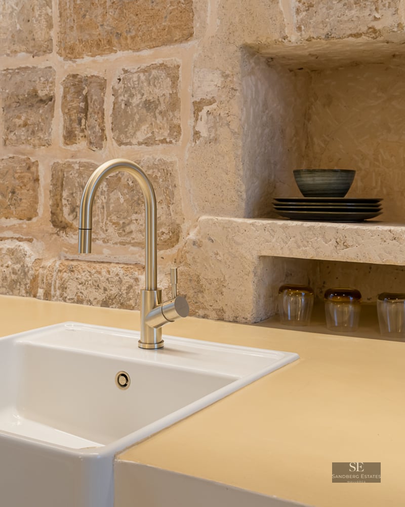 Évier blanc avec robinet doré devant un mur en pierre apparente avec des étagères intégrées dans une cuisine rustique.