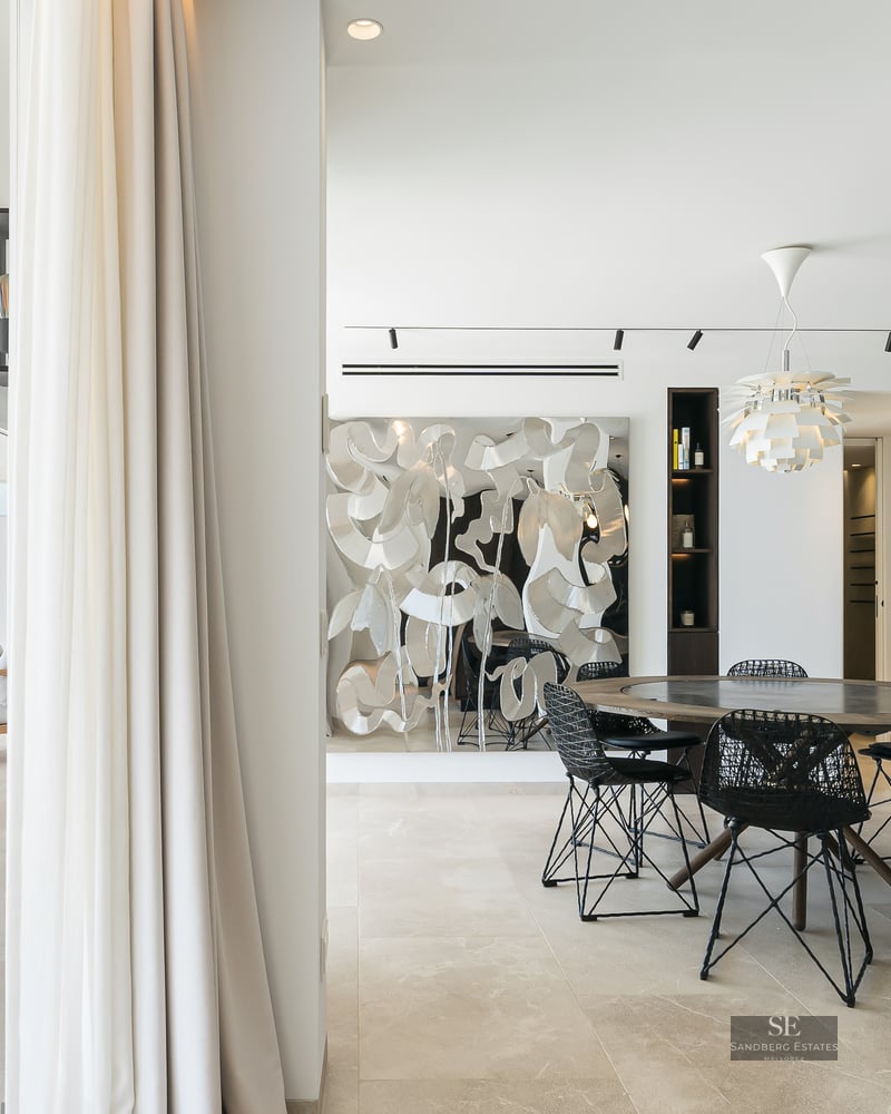 Modern indoor dining area with round wooden table opening onto a bright sun-drenched terrace with lounge seating.