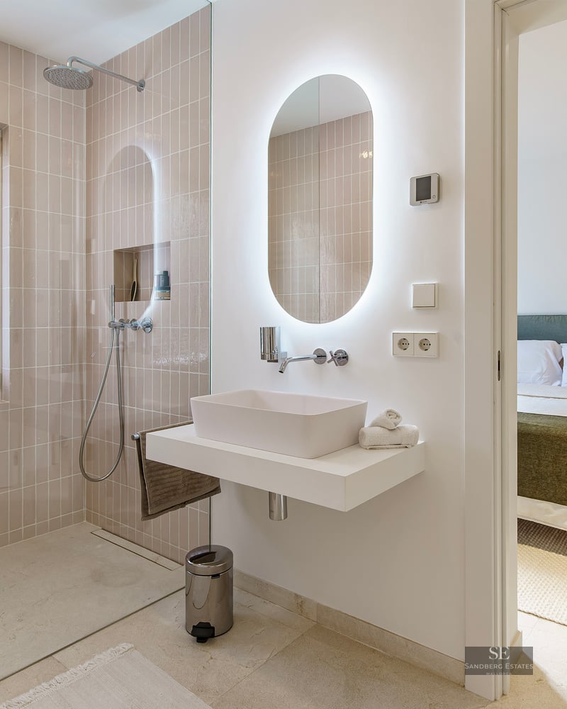 Modern bathroom with walk-in shower, backlit oval mirror, and view into a cozy bedroom.