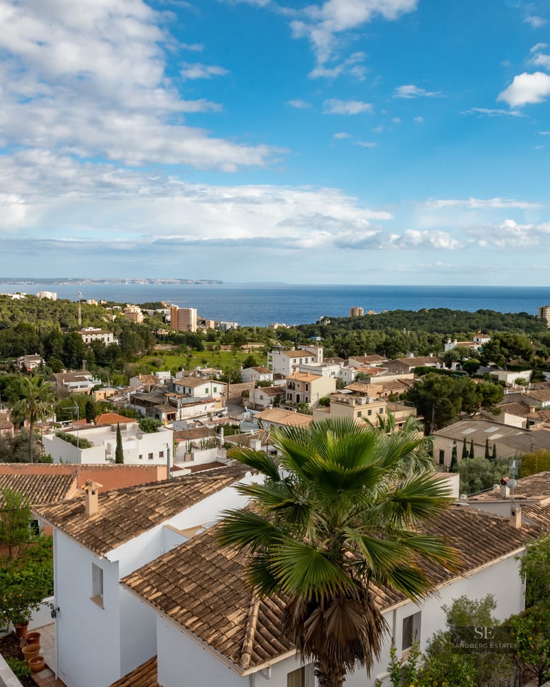 Wide angle view of white villas with terracotta roofs overlooking the blue sea under a partly cloudy sky.