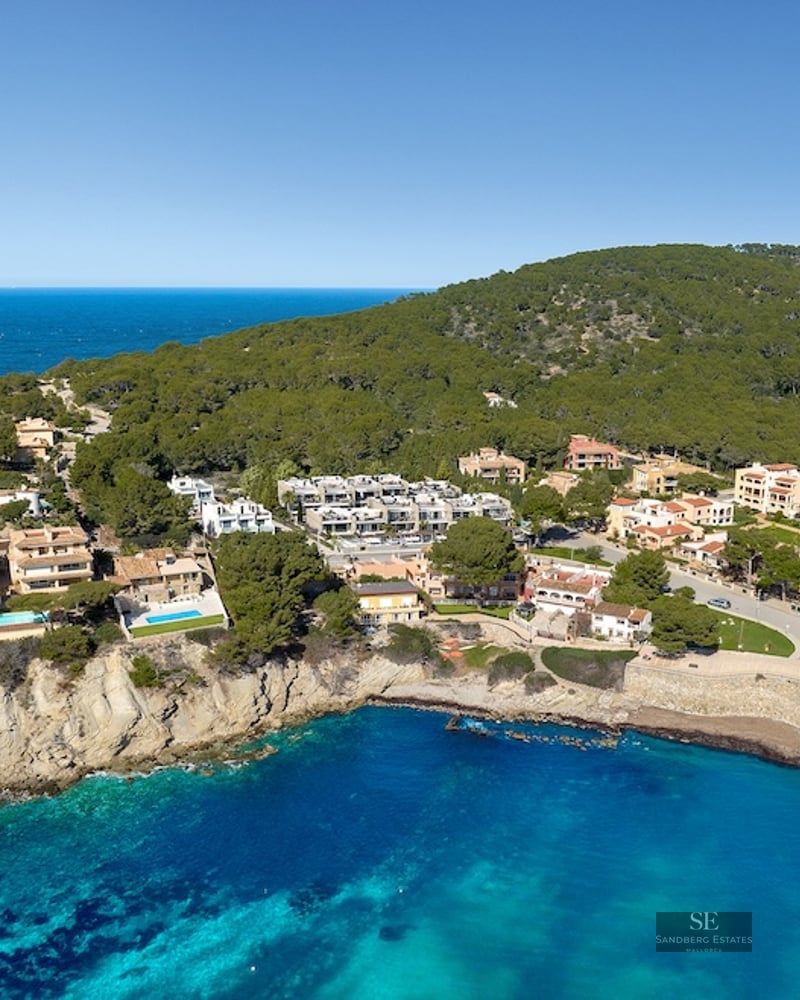 Aerial shot of Mediterranean villas on a rocky cliffside overlooking a turquoise bay and lush green forest.