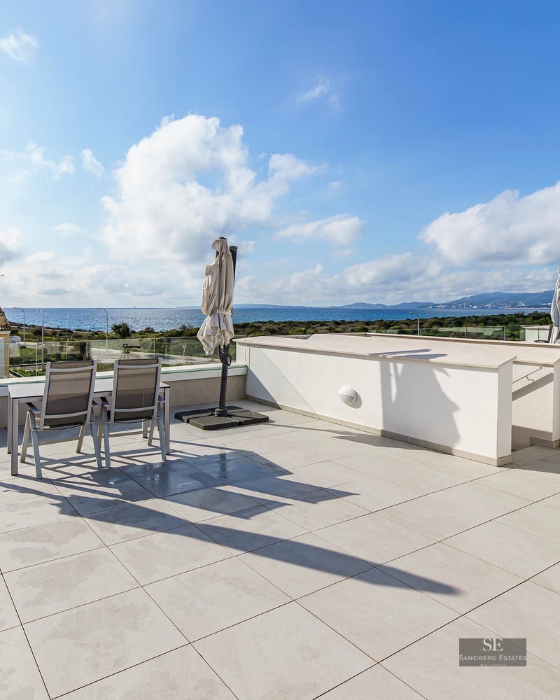 A large modern rooftop terrace with white tiles, glass railings, and a dining set overlooking the blue Mediterranean sea.