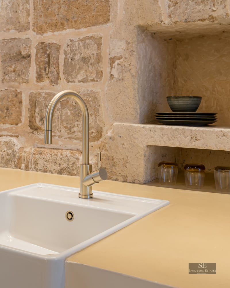 Évier blanc et robinet en laiton devant un mur en pierre naturelle avec des niches de rangement.