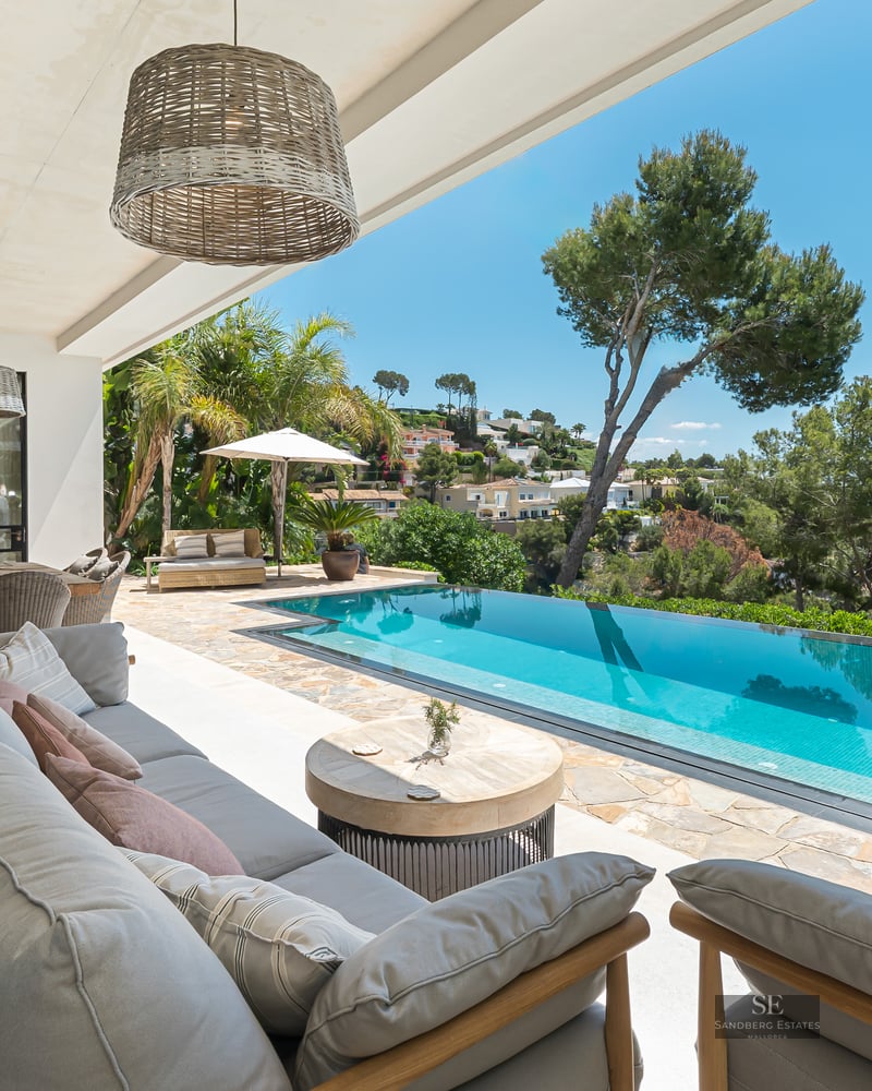 Covered terrace with grey lounge seating overlooking a turquoise infinity pool and the Mediterranean sea.