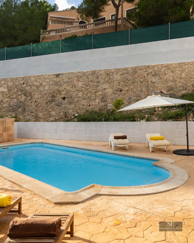 Piscine extérieure avec terrasse en pierre, chaises longues, parasol blanc et mur de pierre.