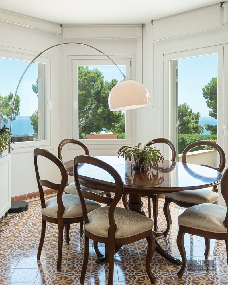 Bright dining room featuring a round wooden table, patterned tile floors, and large windows overlooking the sea.