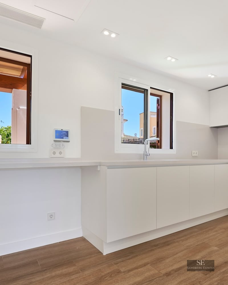 Modern white kitchen with handleless cabinets, wooden floors, and large windows with natural light.
